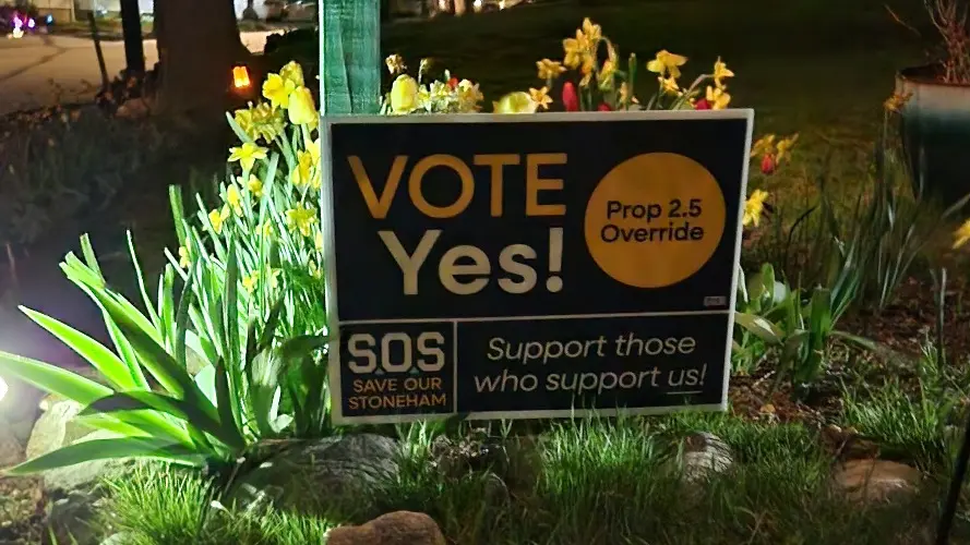 A night shot of a yard sign reading 'Vote Yes' on the Prop 2.5 override in front of a flower bed.