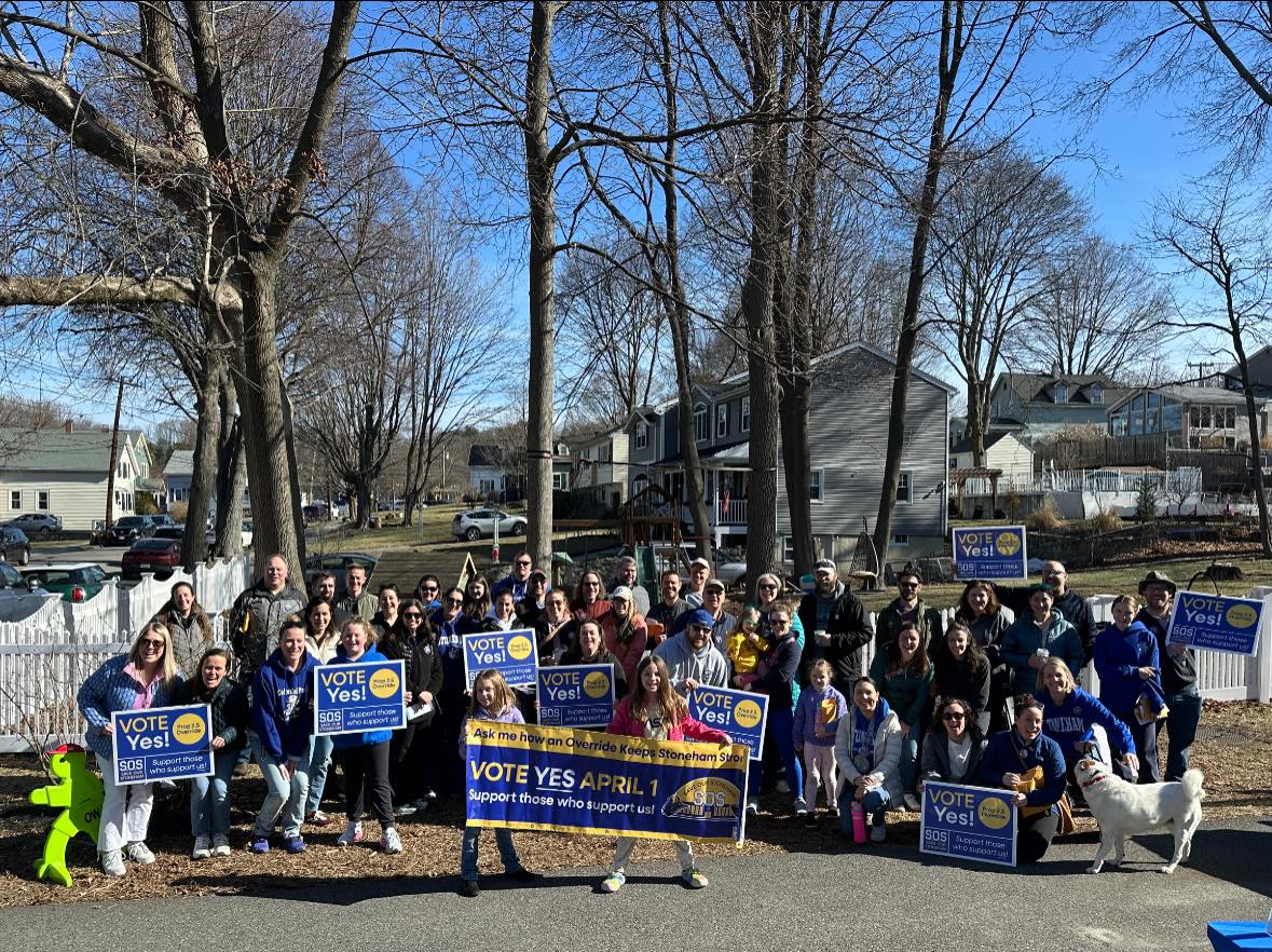Group photo of canvassers in front of a white picket fence holding 'Vote Yes' signs and banners. There is also a very good dog.